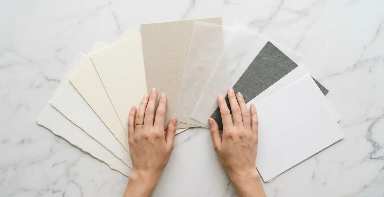 Mains caressant délicatement différentes textures de papiers premium disposés en éventail sur une surface neutre